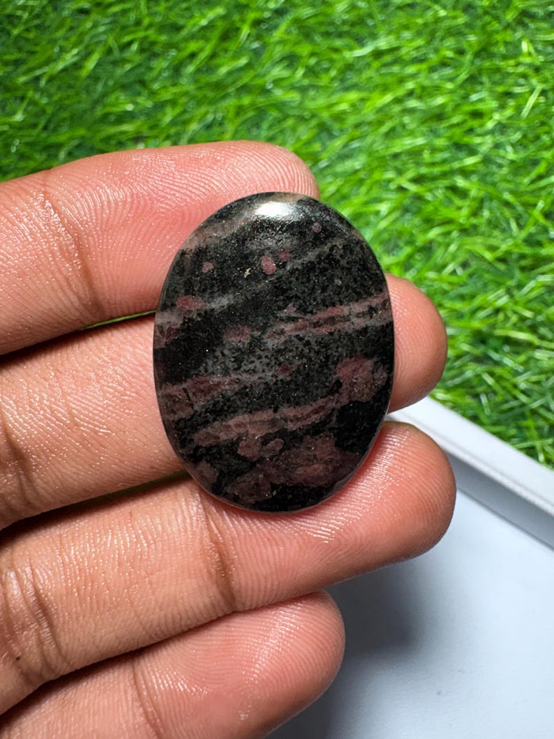 Pu&ograve; includere: Una pietra preziosa lucidata di forma ovale con una base di colore nero scuro e prominenti inclusioni minerali rosso intenso e marrone su tutta la superficie, caratterizzata da una finitura liscia, riflettente e lucida.