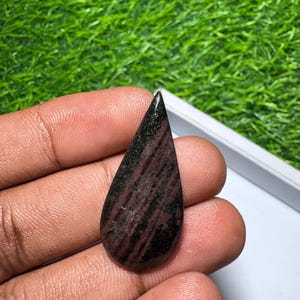May include: A teardrop-shaped gemstone featuring a polished surface with a dark brown and black striped pattern. The stone has a smooth, rounded base that tapers to a sharp point at the top, displaying natural mineral variations throughout.