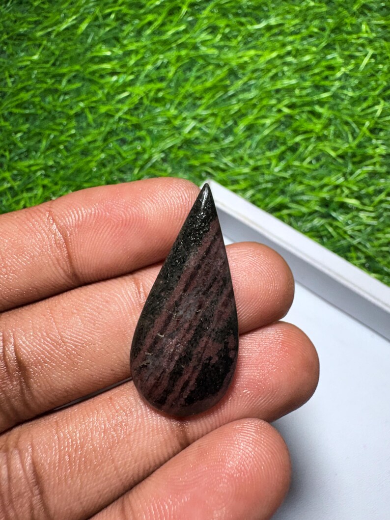 May include: A teardrop-shaped gemstone featuring a polished surface with a dark brown and black striped pattern. The stone has a smooth, rounded base that tapers to a sharp point at the top, displaying natural mineral variations throughout.