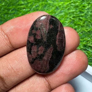 Pode incluir: Uma pedra preciosa polida de formato oval com uma superf&iacute;cie lisa e um padr&atilde;o manchado de preto profundo e bord&ocirc; escuro. A pedra tem um acabamento brilhante que reflete a luz, destacando as bordas arredondadas e a face plana.