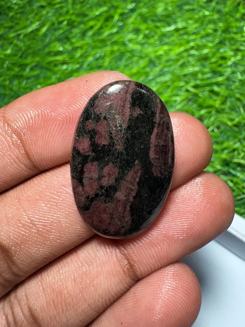 Pode incluir: Uma pedra preciosa polida de formato oval com uma superf&iacute;cie lisa e um padr&atilde;o manchado de preto profundo e bord&ocirc; escuro. A pedra tem um acabamento brilhante que reflete a luz, destacando as bordas arredondadas e a face plana.