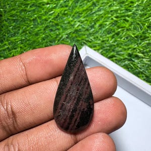 May include: A teardrop-shaped cabochon gemstone featuring a polished surface with deep maroon and black streaks. The stone displays a smooth, rounded finish with a pointed top, showcasing natural color variations in dark red and black tones.