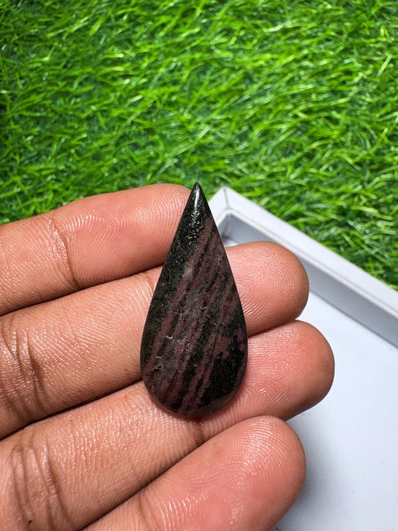 May include: A teardrop-shaped cabochon gemstone featuring a polished surface with deep maroon and black streaks. The stone displays a smooth, rounded finish with a pointed top, showcasing natural color variations in dark red and black tones.