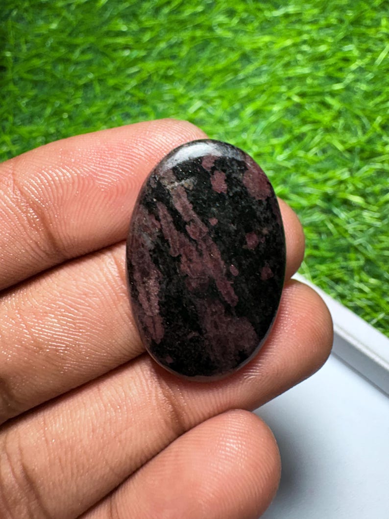 Peut inclure: Une pierre pr&eacute;cieuse cabochon de forme ovale pr&eacute;sentant une surface polie avec un motif tachet&eacute; de couleurs noir profond et rouge bordeaux fonc&eacute;. La pierre a un dessus lisse et arrondi ainsi qu'une finition brillante qui refl&egrave;te la lumi&egrave;re.