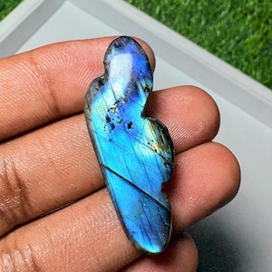 May include: A polished labradorite gemstone with a distinct elongated, irregular shape. The stone displays a vibrant, iridescent blue flash across its surface, contrasted by dark grey inclusions and fine linear fractures throughout the mineral.