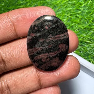 Pu&ograve; includere: Una pietra preziosa lucidata di forma ovale caratterizzata da una base di colore nero scuro con evidenti inclusioni minerali rosso intenso e marrone su tutta la superficie, con una finitura liscia e riflettente e un motivo naturale.