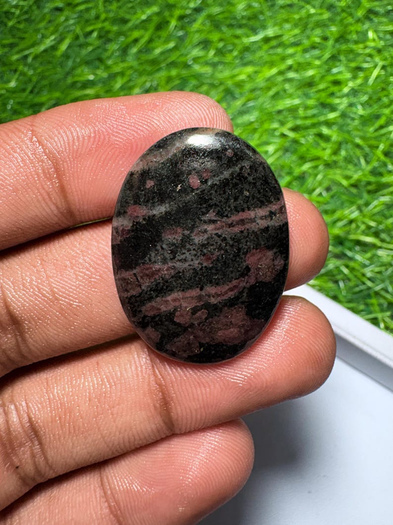 Pu&ograve; includere: Una pietra preziosa lucidata di forma ovale caratterizzata da una base di colore nero scuro con evidenti inclusioni minerali rosso intenso e marrone su tutta la superficie, con una finitura liscia e riflettente e un motivo naturale.