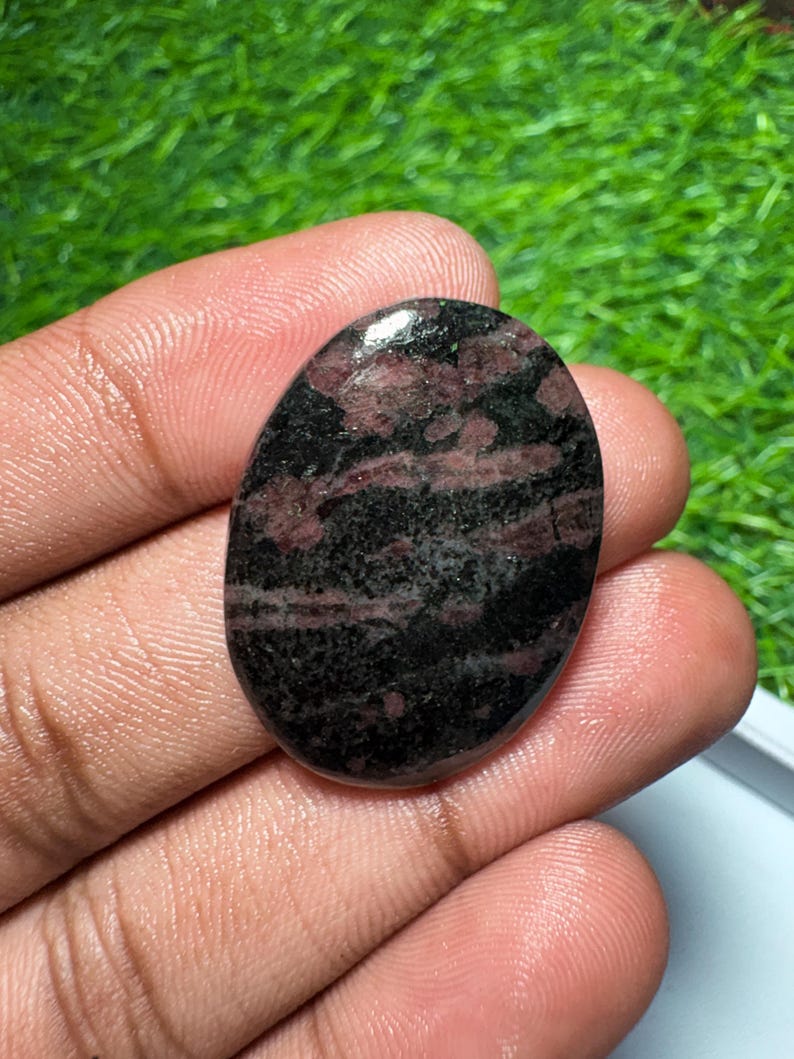 Pu&ograve; includere: Una pietra preziosa lucidata di forma ovale con una superficie liscia e un motivo maculato di colori nero profondo e rosso bordeaux scuro. La pietra ha una finitura lucida che riflette la luce, evidenziando la distribuzione irregolare dei colori.