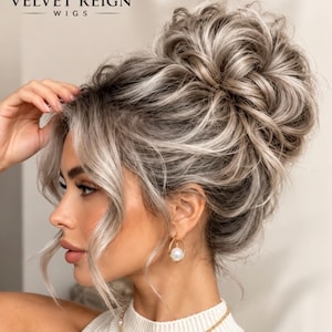 Könnte beinhalten: Eine hohe, voluminöse Hochsteckfrisur mit aschblonden und platinfarbenen Highlights sowie losen, das Gesicht umrahmenden Strähnen. Das Bild enthält den Text Velvet Reign Wigs mit einem Kronenlogo. Die Frisur ist strukturiert und hochgesteckt.
