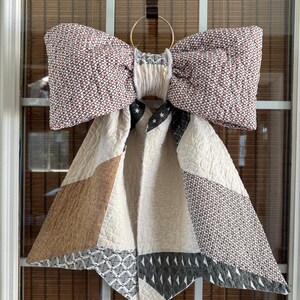 May include: A large fabric bow decoration featuring a quilted texture with a mix of patterns including small geometric shapes, stars, and solid tan panels. The bow is gathered at the center with white fabric and hangs from a gold metal ring.