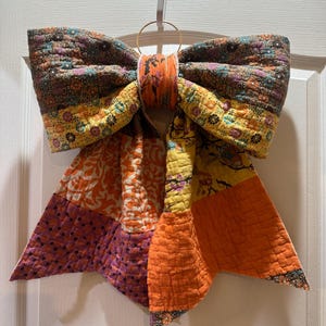 Peut inclure: Un grand nœud décoratif fabriqué à partir de pièces de tissu matelassé aux motifs et couleurs variés, notamment orange, jaune, marron et violet. Le nœud présente un centre noué et deux longs pans pointus, suspendu par un anneau en métal doré.