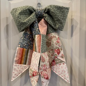 May include: A large decorative bow made from quilted fabric featuring a sage green polka dot pattern at the top. The hanging tails consist of a patchwork design with floral prints, pink roses, vertical stripes, and small bow motifs in muted tones.
