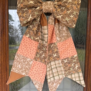 May include: A large decorative bow made from a patchwork of fabric pieces. The design features a mix of brown, tan, and peach colours with patterns including stylised birds, floral prints, and plaid. The bow has two long, pointed tails.