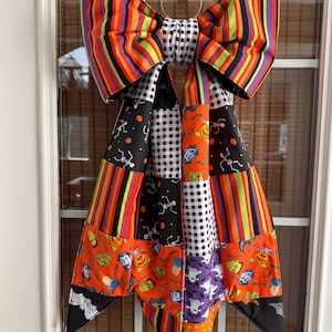 Può includere: Un grande fiocco decorativo in tessuto con un mix di motivi a tema Halloween, tra cui strisce arancioni, nere, viola e verdi, quadretti vichy bianchi e neri, scheletri, zucche, fantasmi e vari volti di mostri, appeso a un anello in metallo.