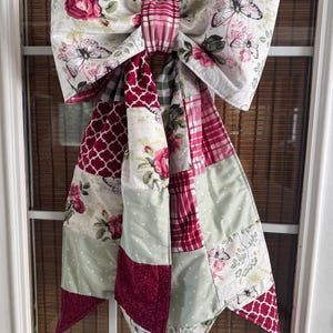 Könnte beinhalten: Eine große Stoffschleife mit Patchwork-Design und verschiedenen Mustern, darunter rote Rosen, schwarze Schmetterlinge, weiße Punkte und geometrische Gitterdrucke in Rot-, Salbeigrün- und Weißtönen, aufgehängt an einem goldfarbenen Metallring.