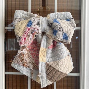 May include: A large decorative bow constructed from quilted fabric patches in various colors including light blue, cream, brown, and red. The fabric features frayed edges and a stitched grid pattern throughout. The bow is suspended from a gold metal ring.
