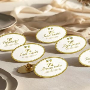 May include: A collection of oval-shaped metal name badges with gold-coloured borders and white centres. Each badge features a small green leaf icon and text including The Papparazzi, The Toast master, The Hype person, The Tissue provider, and The Memory maker.