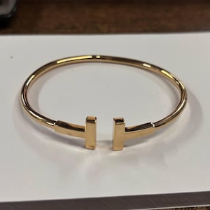 May include: A polished gold-tone metal cuff bracelet featuring a circular band that remains open at the front. The two ends of the bracelet terminate in flat, T-shaped bars that face each other, creating a distinct gap in the center of the design.