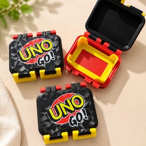 May include: Two black plastic UNO GO! card game cases with geometric patterns and yellow latches, one closed and one open to reveal a red interior, alongside a third closed case featuring the same black, yellow, and red color scheme.