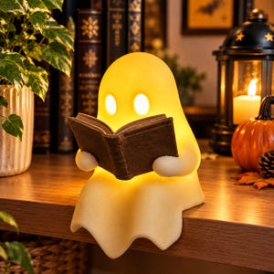 May include: A glowing, cream-colored ghost-shaped lamp sitting on a wooden surface. The ghost has two oval-shaped eyes and is holding an open, dark brown book in its hands. The lamp emits a warm, soft light from within its smooth, rounded body.