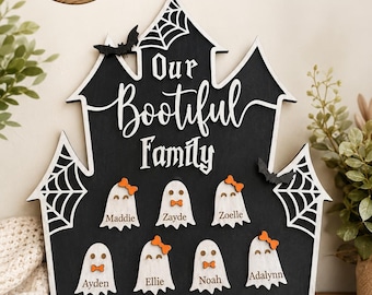 Puede incluir: Un letrero de madera negro con forma de casa encantada, detalles de telarañas blancas y dos murciélagos negros. El texto dice Our Bootiful Family. Debajo hay siete figuras de fantasmas blancos con lazos o corbatines naranjas y nombres.