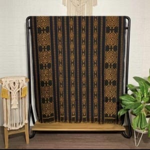 Puede incluir: Un textil rectangular con fondo negro, rayas verticales y patrones geométricos repetitivos en un tono marrón dorado. La tela está colocada sobre un marco de metal negro y presenta un borde inferior con flecos y un diseño tejido.