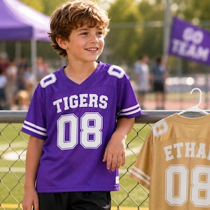 May include: A purple V-neck jersey with white text reading TIGERS and the number 08 on the front, and a gold jersey with white text reading ETHAN and the number 08 on the back, both featuring white stripes on the sleeves and shoulder numbers.