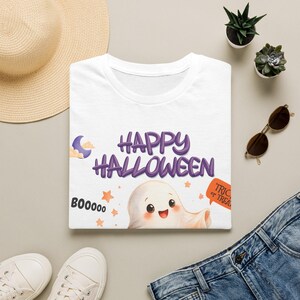 Puede incluir: Una camiseta blanca con un gráfico morado de Happy Halloween, un fantasma sonriente, estrellas naranjas, una luna creciente, el texto BOOOOO y un bocadillo naranja que dice TRICK OR TREAT, centrado en la parte frontal de la prenda.