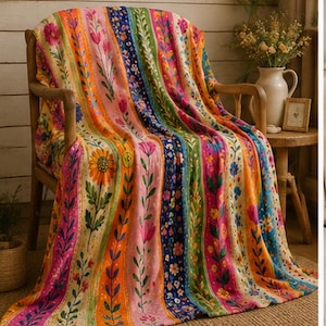 May include: A soft throw blanket featuring a vibrant vertical striped pattern with various floral motifs in shades of orange, pink, green, blue, and yellow. The blanket is draped over a wooden chair, displaying detailed illustrations of sunflowers, tulips, and small blossoms.