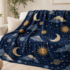 May include: A dark blue fleece throw blanket featuring a celestial pattern of golden suns with faces, crescent moons with faces, planets with rings, and swirling gray clouds interspersed with small yellow stars and constellations.