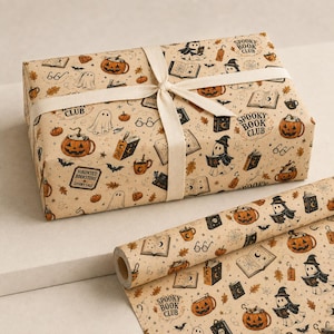 May include: A rectangular gift box wrapped in beige paper with a repeating pattern of ghosts, pumpkins, books, and the text Spooky Book Club, tied with a cream ribbon. A matching roll of the same patterned wrapping paper lies in the foreground.