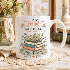 May include: A white ceramic mug featuring a stack of three books topped with colorful flowers. The text reads "NEVER Forget the DIFFERENCE YOU MAKE" and "Mrs. Martin" on a green banner. Small hearts and floral accents decorate the design.