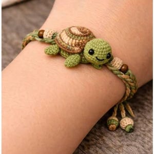 May include: A bracelet featuring a small, crocheted green turtle with a brown patterned shell. The band is made of braided green and tan cord, accented with small brown and tan beads. Two dangling cord ends are finished with small crocheted beads.