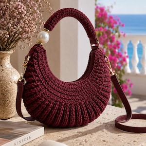 Puede incluir: Un bolso de ganchillo de color burdeos oscuro en forma de media luna, con un asa gruesa y redondeada acentuada por una sola perla blanca artificial grande. El bolso incluye una correa de hombro a juego desmontable con herrajes metálicos dorados.