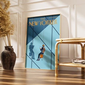 May include: A framed poster of The New Yorker magazine cover dated July 7 & 14, 2025. The illustration features two window washers suspended by ropes against a blue geometric building facade, with one worker wearing an orange helmet and bucket.