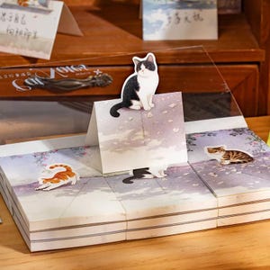 May include: A set of adhesive sticky notes featuring illustrations of cats. The notes display a black and white cat sitting on a roof, an orange tabby cat stretching, and a brown tabby cat resting on a path with floral accents and soft colors.