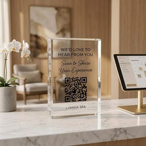 May include: A clear rectangular acrylic sign featuring black text that reads WE'D LOVE TO HEAR FROM YOU, Scan to Share Your Experience, and LUMINA SPA, along with a central black QR code. The sign is positioned upright on a white marble surface.