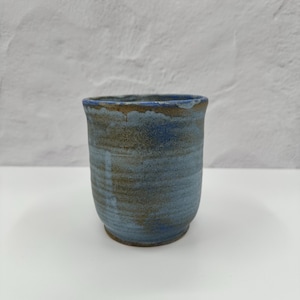May include: A cylindrical ceramic cup featuring a mottled glaze in shades of blue and earthy brown. The surface displays a textured, hand-thrown appearance with subtle horizontal ridges and a slightly uneven rim, showcasing a blend of cool and warm tones.
