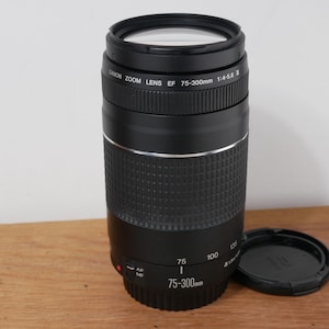 Może przedstawiać: Czarny obiektyw Canon Zoom Lens EF 75-300mm 1:4-5.6 III z teksturowanym gumowym pierścieniem ostrości, srebrnym pierścieniem ozdobnym i oddzielną czarną pokrywką. Na korpusie znajduje się biały nadruk z parametrami ogniskowej i przysłony.