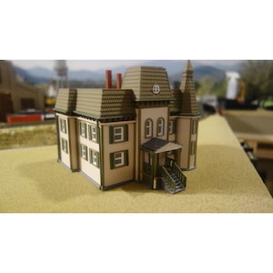 Può includere: Un modellino in miniatura di una casa a due piani con rivestimento beige, persiane verde scuro e un tetto a mansarda verde scuro. La struttura presenta un portico con scale, due camini rossi, una piccola torre e diverse finestre rettangolari.