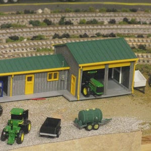 Könnte beinhalten: Ein Miniatur-Bauernhofgebäude mit grauer vertikaler Fassade, grünem Dach und gelben Verzierungen an Türen und Fenstern. Das Gebäude verfügt über eine offene Bucht mit einem grünen Traktor sowie weitere Miniatur-Traktoren und Anhänger.