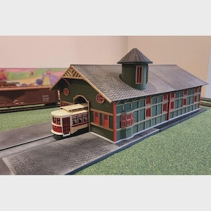 Könnte beinhalten: Ein Modell-Bahnhofsgebäude mit dunkelgrünen Wänden, roten Fensterrahmen und einem grauen Schindeldach, mit einem kleinen Turm und einer cremefarbenen und roten Straßenbahn, die aus dem Seiteneingang kommt, auf einer grünen Basis mit schwarzen Gleisen.