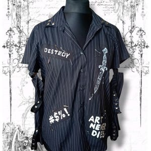 Könnte beinhalten: Ein schwarzes kurzärmeliges Hemd mit weißen Nadelstreifen, verziert mit Aufnähern mit der Aufschrift "DESTROY", "#$%!" und "ART NEVER DIES", einem weißen Dolchmotiv, Sicherheitsnadeln sowie Metallnieten am Kragen und an den Ärmeln.