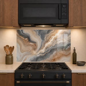 May include: A rectangular kitchen backsplash panel featuring a swirling abstract pattern of gray, beige, and white tones with metallic gold accents. The panel is positioned behind a black gas stove and below a black microwave oven in a kitchen.