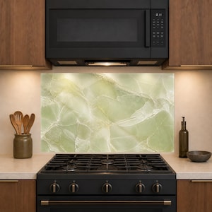 May include: A rectangular kitchen backsplash panel featuring a light green marble pattern with white veining. The panel is positioned between a black microwave and a black gas stove, with wooden cabinets above and a countertop below.