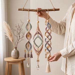 May include: Three hanging macrame ornaments suspended from a horizontal wooden branch. Each piece features intricate crochet patterns in muted tones of blue, pink, and cream, accented with wooden beads, gold star or snowflake charms, and clear crystals.