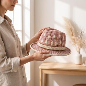 May include: A crocheted fedora hat in dusty rose and cream, featuring a repeating diamond pattern on the crown and a thick, textured cream-colored band above the brim. The hat is constructed with a dense, uniform stitch throughout the design.