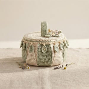 May include: A round, lidded crochet basket featuring alternating panels of cream and sage green yarn. The lid is adorned with small crocheted flowers, while the rim is decorated with a string of beads and dangling, leaf-shaped crochet accents.
