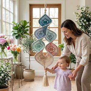 May include: A spiral-shaped crochet hanging decoration featuring a gradient of colors including teal, light blue, sage green, dusty rose, and cream. The piece is suspended from a cord with a wooden bead and ends with a matching cream-colored tassel.