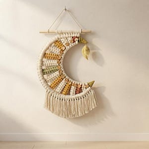 May include: A crescent moon-shaped macrame wall hanging made of off-white cord, featuring alternating sections of mustard yellow, sage green, and terracotta knotted patterns. Small wooden buttons are attached along the curve, with a fabric leaf.